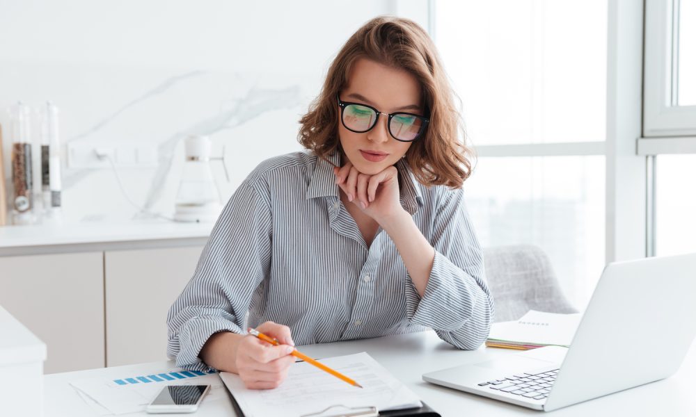 Young concentrated businesswoman in glasses and striped shirt working with papers at home